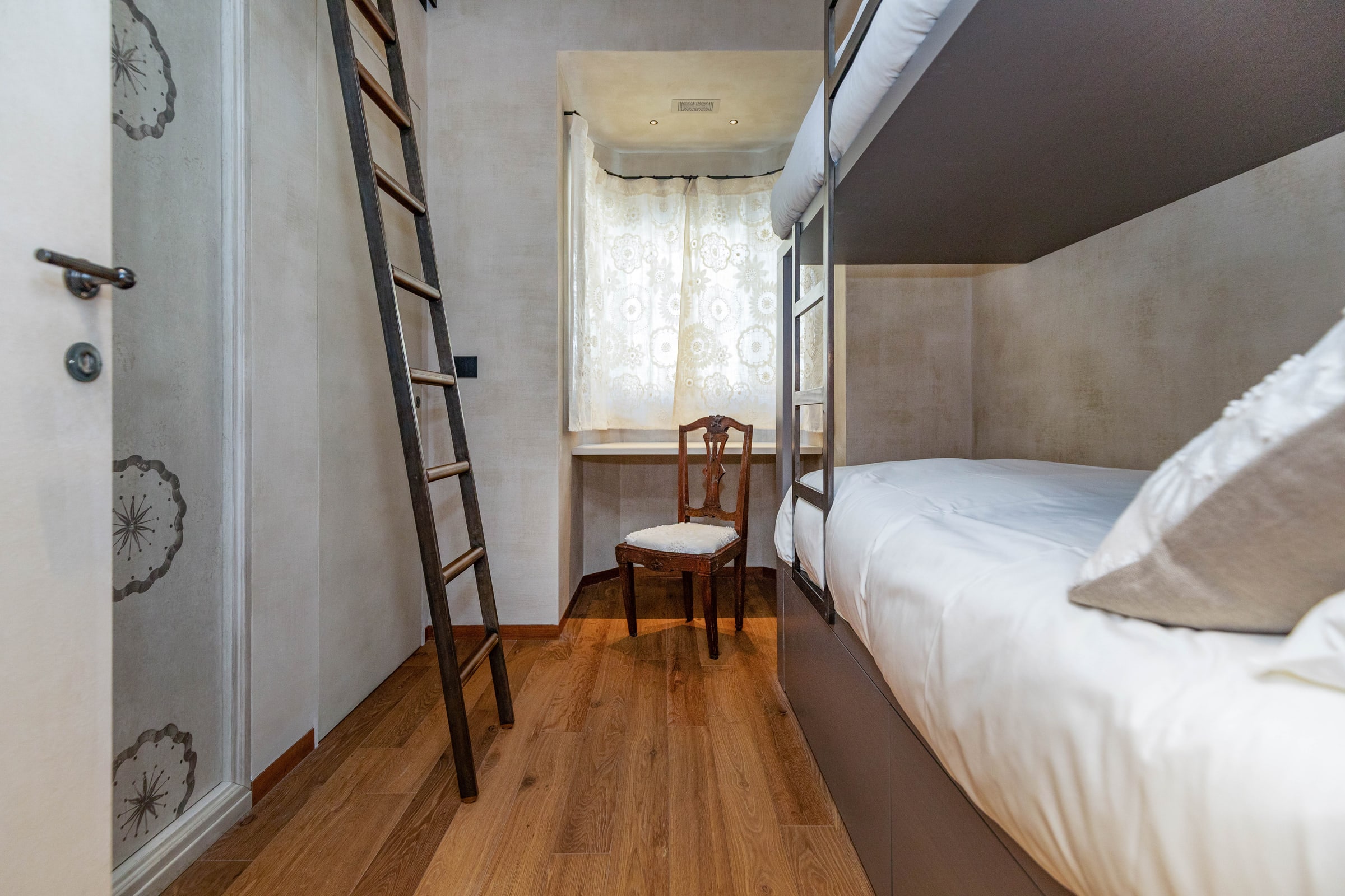 Camera con letto a castello, scala in metallo, finestra con tende e sedia tirolese dipinta