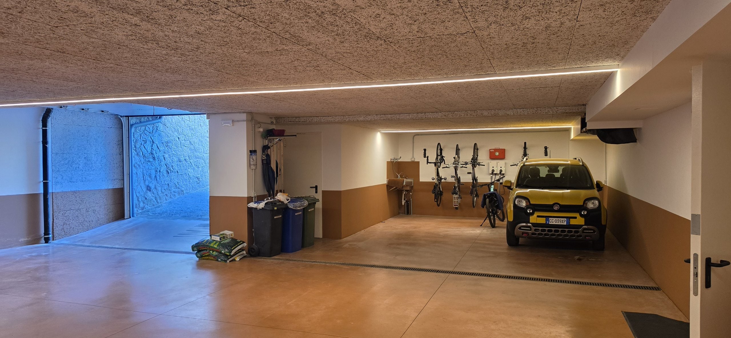 Garage with the door open to daylight, a small yellow car parked and skis and bicycles on the wall racks