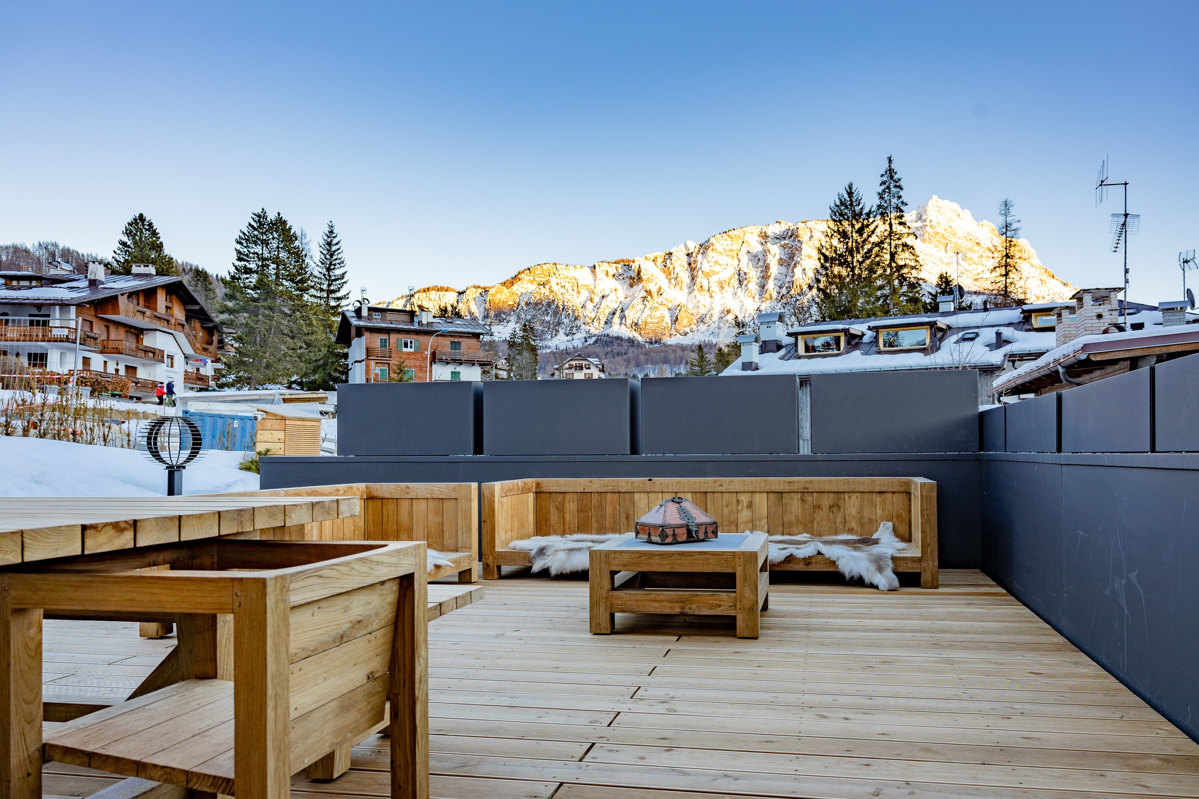 Wooden sun terrace with benches and a low table, the snow-covered roofs of Cortina and the Dolomites lit at sunset