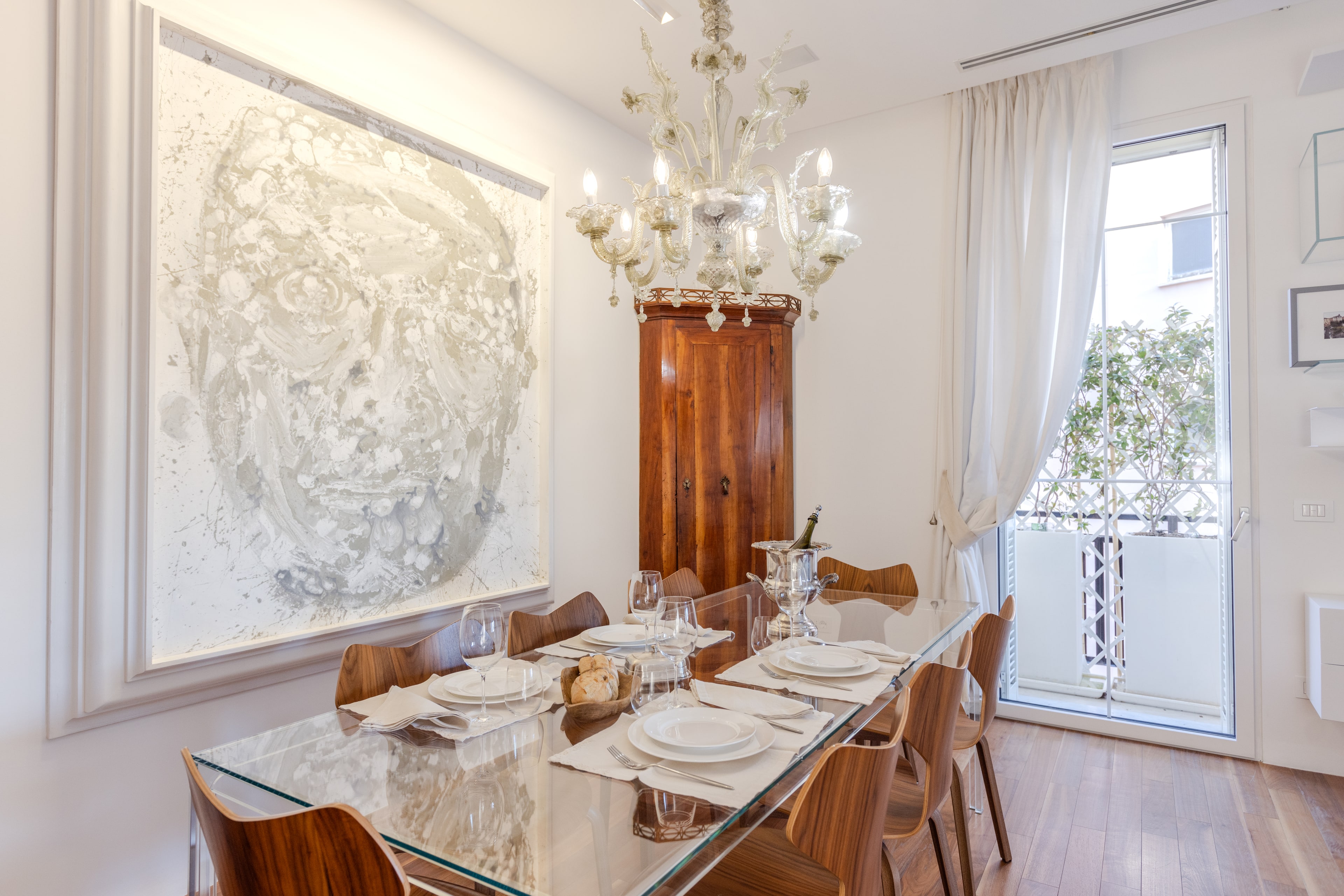 Sala da pranzo con grande ritratto di perle, lampadario di Murano, tavolo di cristallo apparecchiato e cabinet in noce