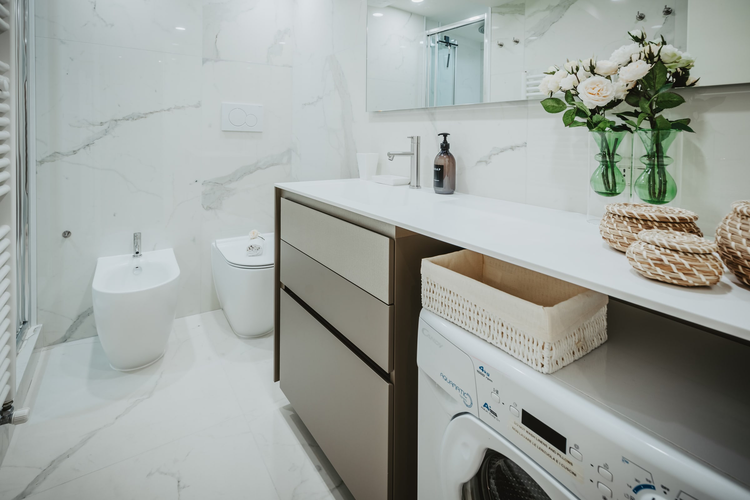Bagno luminoso in marmo bianco con bidet, vasi di rose bianche, doccia in vetro e mobile lavabo chiaro